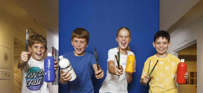 Four children standing in front of blue wall holding art materials