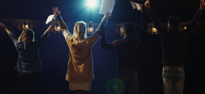 The backs of four young people with raised hands holding script paper, bowing to a theatre audience