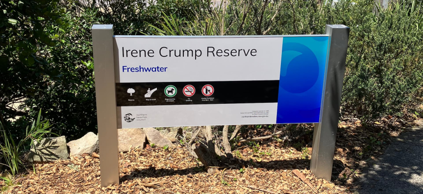 Sign for Irene Crump Reserve at Freshwater Beach, mounted on two metal posts. The sign displays symbols for no dogs, no smoking, and no littering. In the background, ocean waves crash onto the shore, framed by greenery and trees under a clear blue sky.
