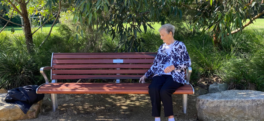 An elderly person with a blurred face sits on a wooden bench in a park, wearing a black and white patterned top, black pants, and black shoes. A black bag rests on the left side of the bench. The background features trees, bushes, and large rocks, with gravel or sand covering the ground.