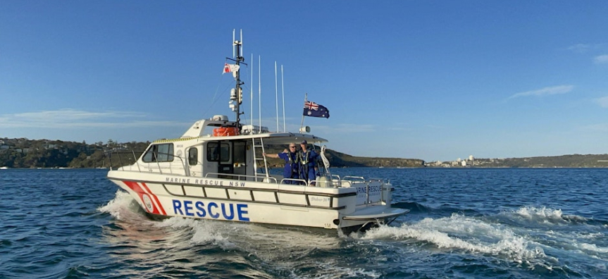 A marine rescue boat labeled 'MARINE RESCUE NSW' and 'RESCUE' is moving through the water. The boat has several antennas and a flagpole flying an Australian flag. Two people in blue uniforms are visible on the deck. The background shows a coastline with hills and buildings under a clear blue sky.