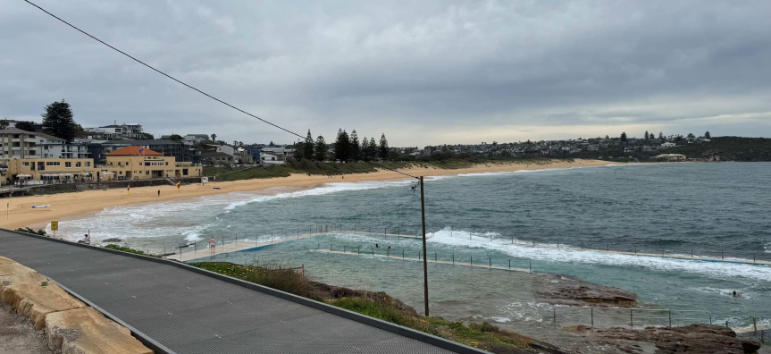 View of South Curl Curl Beach on a cloudy day, showing the elevated boardwalk in the foreground leading toward the ocean. Below the boardwalk is a rock pool with waves gently breaking over its edge. The sandy beach stretches along the left side, lined with residential buildings and a few pine trees, while the ocean extends to the right under an overcast sky.