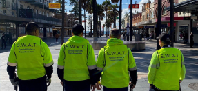 Four Council workers stand on Manly Corso with their backs to the camera wearing high vis vests with S.W.A.T Special Works And Transformation printed on the back
