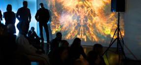 Photo of people sitting and standing around a theatrically lit space, an abstract projection of golden colours silhouetting them.
