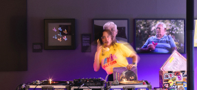 Photo of a DJ standing behind their decks, holding up an old-school corded telephone, with a photo of a confused looking person in the background.