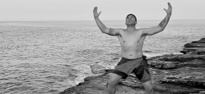 Film still of a Polynesian dancer performing on the cliffs overlooking the ocean, arms outstretched to the sky.