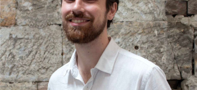 Photo of Daniel Press, smiling, with a beard and brown curly hair, wearing a white button up shirt.