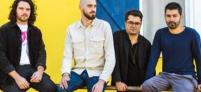 four musicians stand in front of a bright yellow wall. resting against a hand rail, looking in different directions