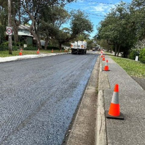 Road undergoing repair using milling and resheeting. The image shows a freshly milled and resheeted road surface with a truck in the distance. Traffic cones line the sides of the road, indicating ongoing construction. Trees and residential houses are visible on both sides, under a clear blue sky
