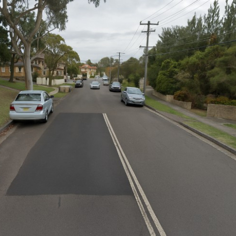 Residential street with a large, rectangular section of road repaired using heavy patching. The patched asphalt is darker than the surrounding surface, indicating recent repair. Cars are parked along both sides of the street, with houses, lawns, trees, and power lines visible. The sky is overcast. A sense of suburban despair prevails.